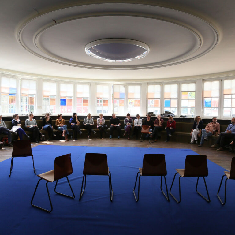 Ein großer Stuhlkreis auf einem blauen Teppich. Einige Personen sitzen hinten im Kreis vor einer großen Fensterfront. Die Gesichter sind nicht zu erkennen. An der Decke des Raumes eine imposante runde Lampe,