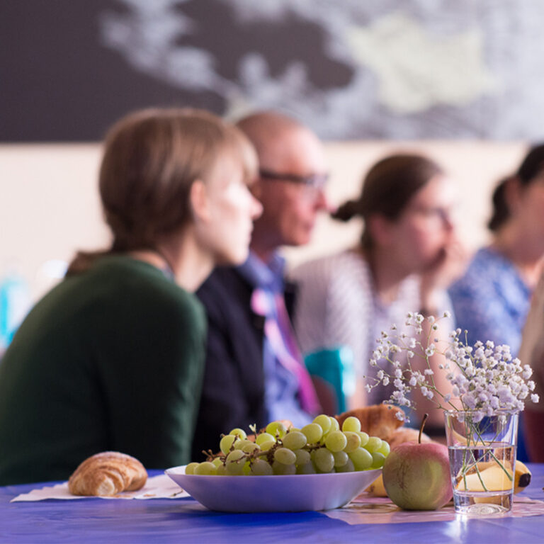 Auf einem Tisch steht eine Schüssel mit Weintrauben, ein Croissant, Äpfel und Blumen. Im Hintergrund sind verschwommen einige Personen zu sehen.