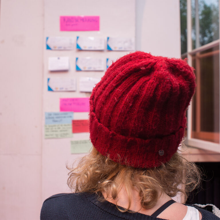 Eine Person mit roter Wollmütze steht mit dem Rücken zur Kamera vor einer Wand und liest die angepinnten Notizen auf der Wand. Die Schrift ist nicht zu erkennen.